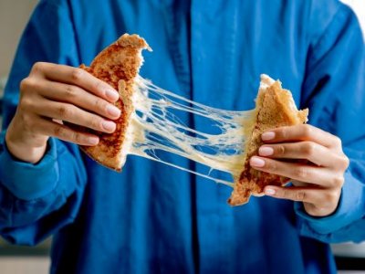 A person in a blue button-down shirt is pulling apart a toasted cheese sandwich, with strings of melted cheese between the two sides.